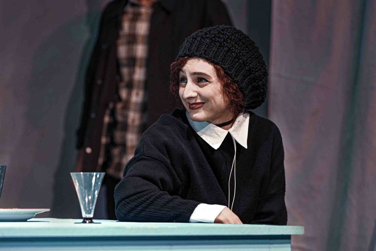 Aida in a dark costume at a table on stage
